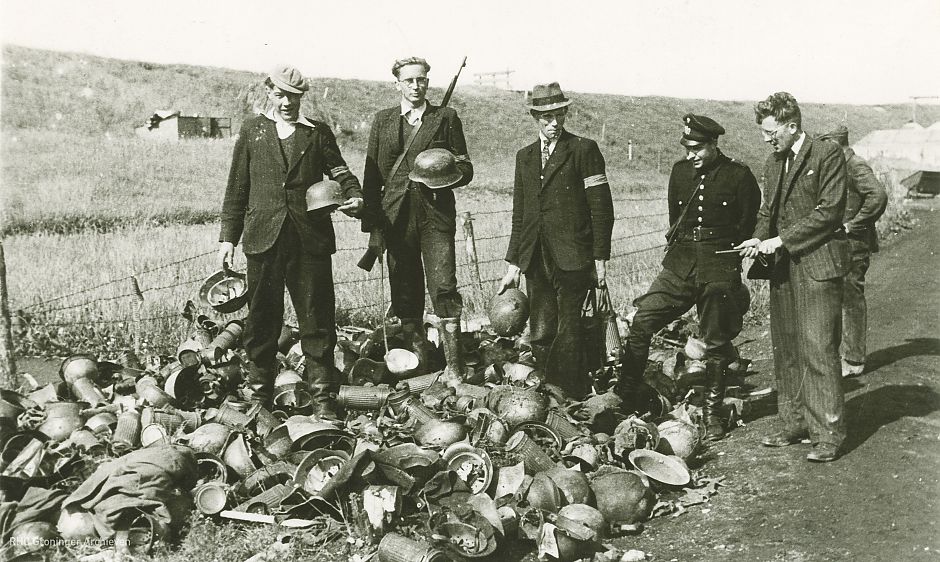 Mannen bij achtergelaten helmen en andere rommel bij het geschut de Fiemel bij Termunten, mei 1945. - Foto: www.beeldbankgroningen.nl (2138-5895)