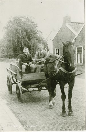 <p>De Hoofdstraat in Finsterwolde: een man en een jongen zitten op de bok van een een paard en wagen geladen met melkbussen, 1955. &ndash; Foto: collectie Groninger Archieven</p>
